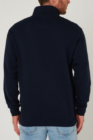 A man is seen from the back wearing a dark navy blue Pilbara Classic Fleece Pullover with a high collar and light blue jeans. He stands against a plain light gray background that highlights the pullover’s fleece lining detail.