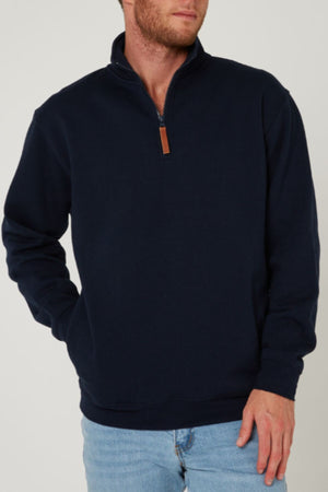 A man wears a navy blue Pilbara Classic Fleece Pullover with cozy fleece lining and light blue jeans, standing with one hand in his pocket against a plain light background.