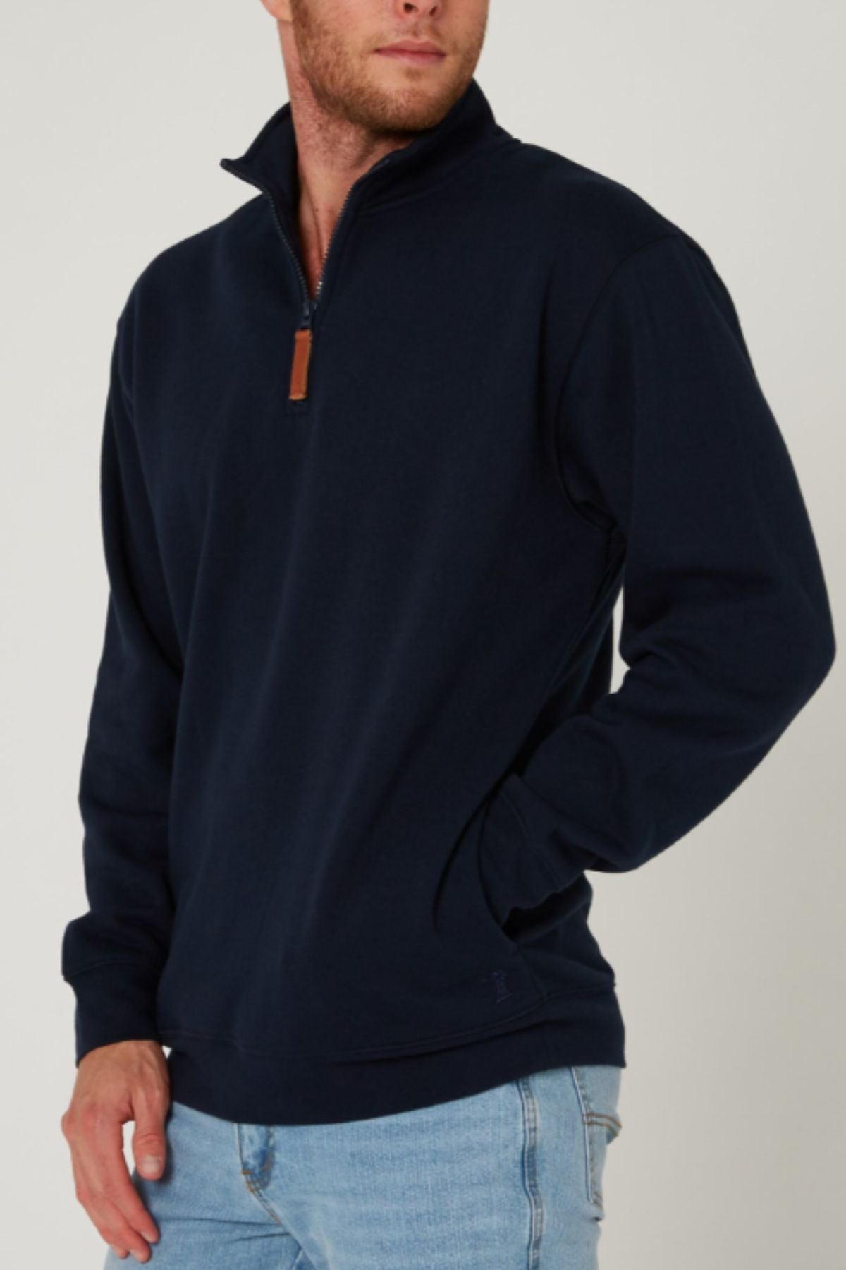 A man wearing a navy blue Pilbara Classic Fleece Pullover with cozy fleece lining and light blue jeans stands with one hand in his pocket against a plain light background.