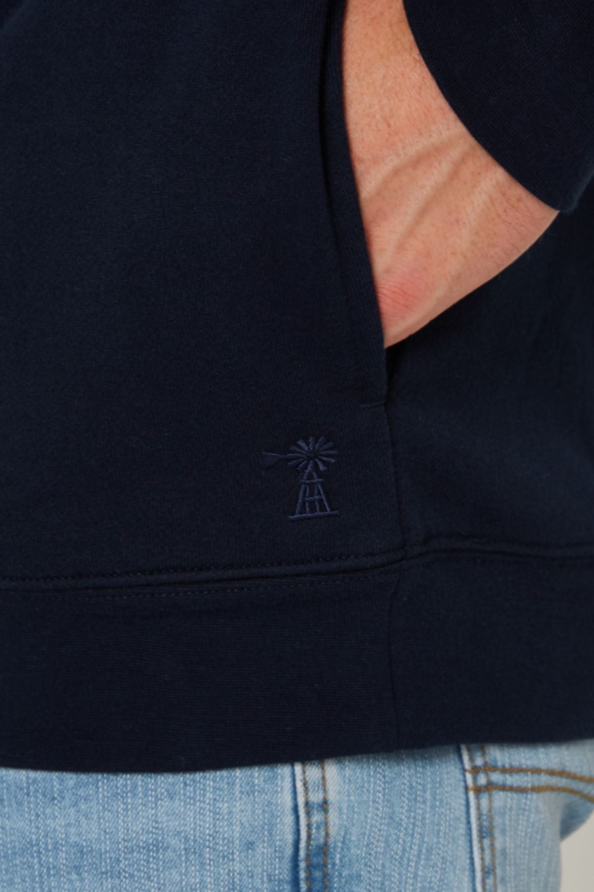 Wearing the Pilbara Classic Fleece Pullover in dark navy with an embroidered windmill logo near the hem and light blue jeans, a person stands with one hand in their pocket.