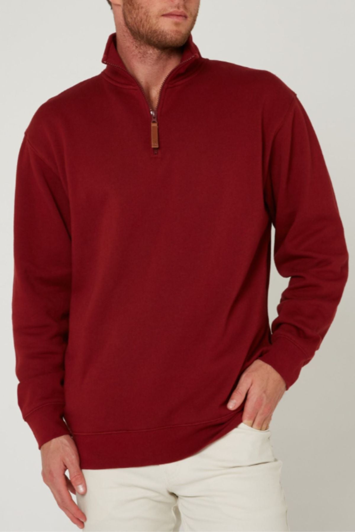 A man stands against a plain light background wearing a maroon Pilbara Classic Fleece Pullover with a high collar, paired with beige pants. His left hand is on his hip and his right hand rests by his side.