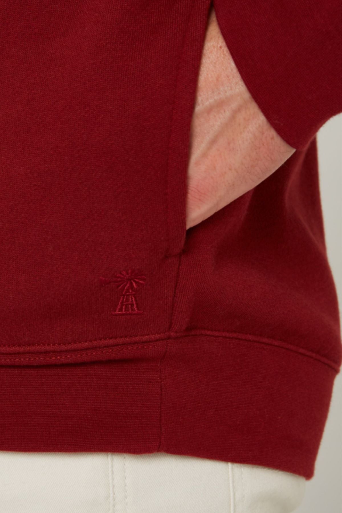 A close-up of a person’s hand in the pocket of a red Pilbara Classic Fleece Pullover with a small embroidered logo near the hem, paired with white pants.