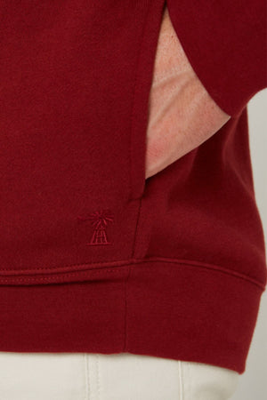A close-up of a person’s hand in the pocket of a red Pilbara Classic Fleece Pullover with a small embroidered logo near the hem, paired with white pants.