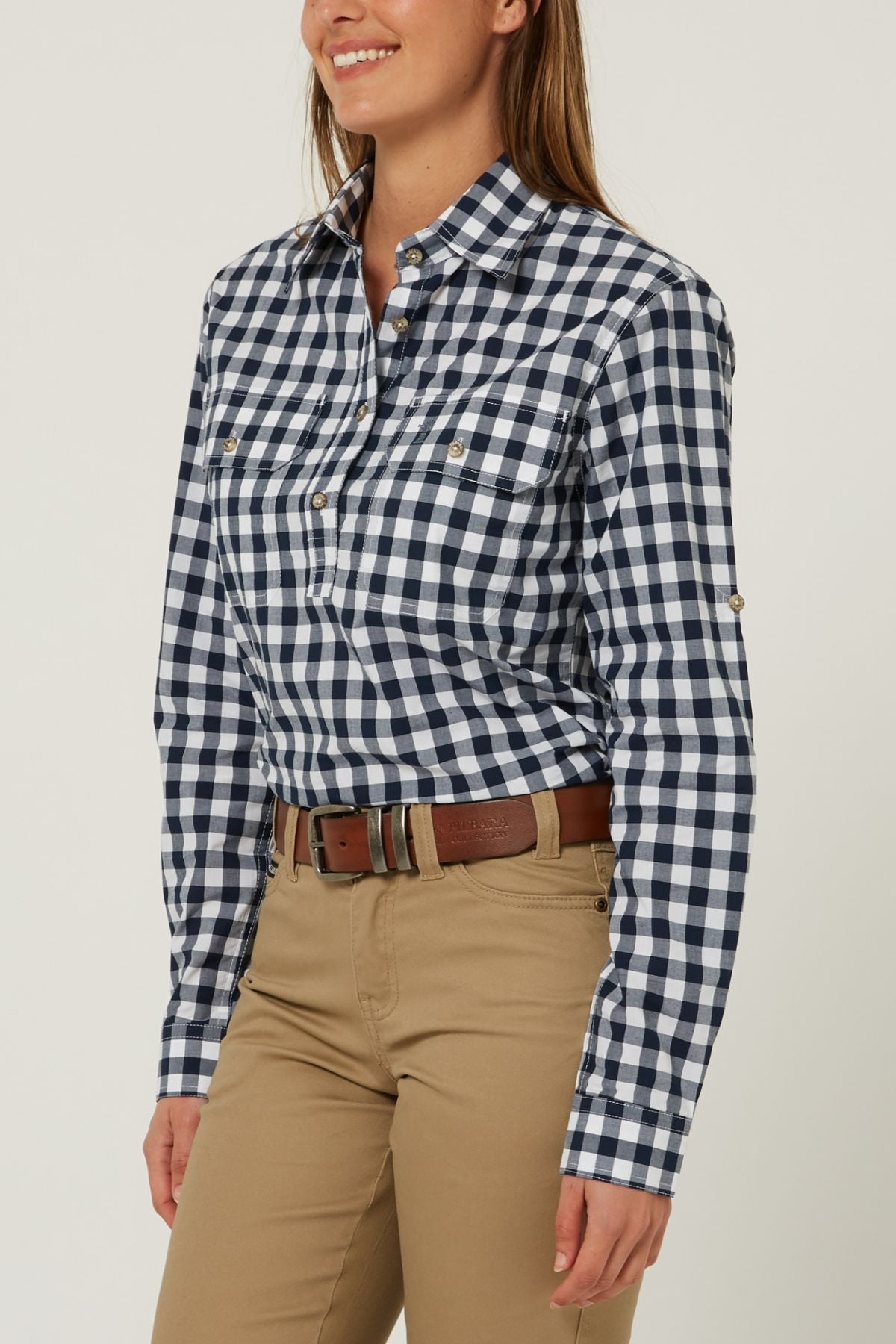 A woman smiles against a light background, wearing the Pilbara Ladies Check Closed Front Long Sleeve Shirt in navy and white, tucked into tan pants with a brown belt. Her long hair is down.