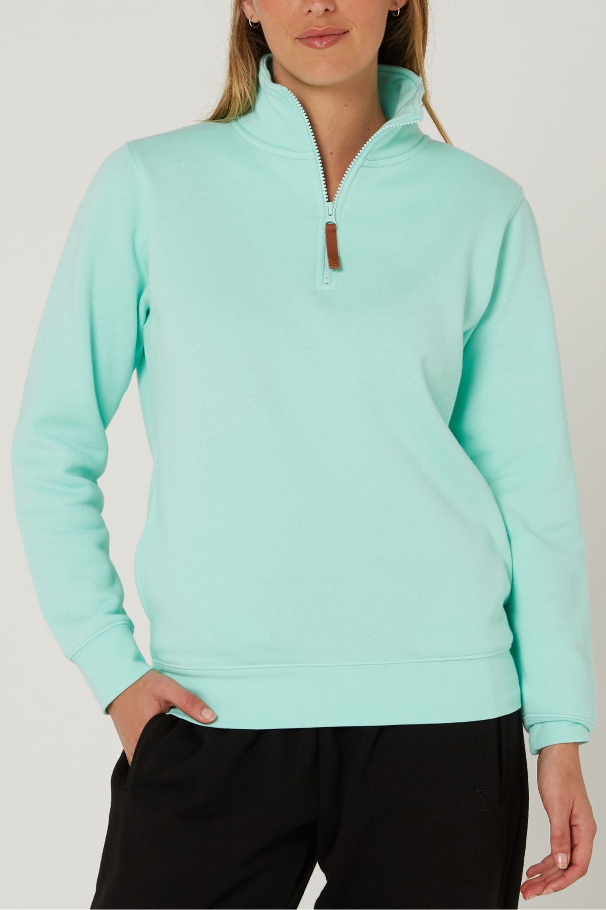 A woman wears the Pilbara Ladies Classic Fleece Pullover in light turquoise with cozy fleece lining and black pants, standing against a plain background with one hand in her pocket.