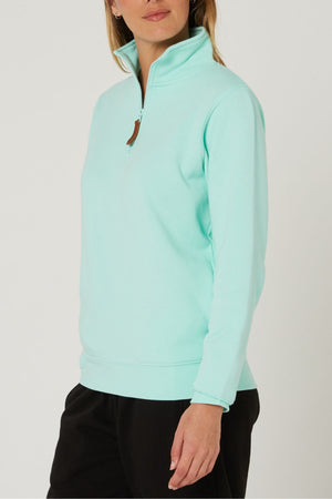 A woman wears the Pilbara Ladies Classic Fleece Pullover in light turquoise with a high collar and cozy fleece lining, paired with black pants, standing against a plain light background.