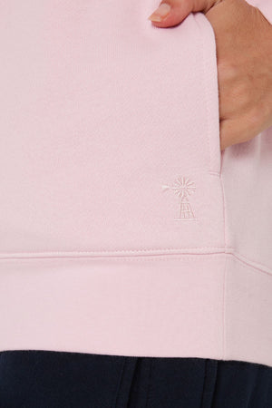 Close-up of a person’s hand in the pocket of a light pink Pilbara Ladies Classic Fleece Pullover with a small embroidered windmill near the hem, paired with dark pants.