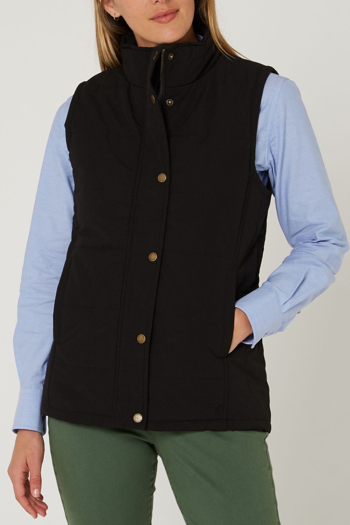 A person wearing the Pilbara Ladies Vest in black with Polyfill padding over a light blue collared shirt and olive green pants stands with one hand in their pocket against a plain background.