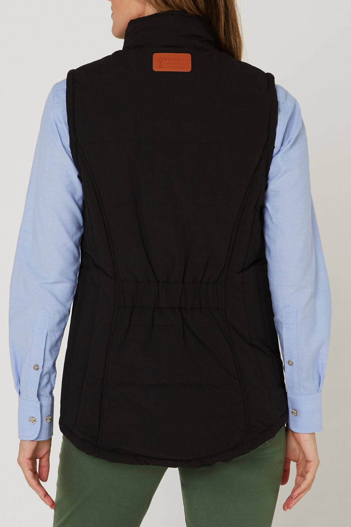 A woman is seen from the back wearing green pants, a light blue long-sleeve shirt, and the Pilbara Ladies Vest—a black winter vest with Polyfill padding and a brown rectangular patch near the collar.