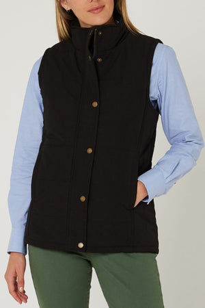 A person wearing the Pilbara Ladies Vest in black with Polyfill padding over a light blue collared shirt and olive green pants stands with one hand in their pocket against a plain background.
