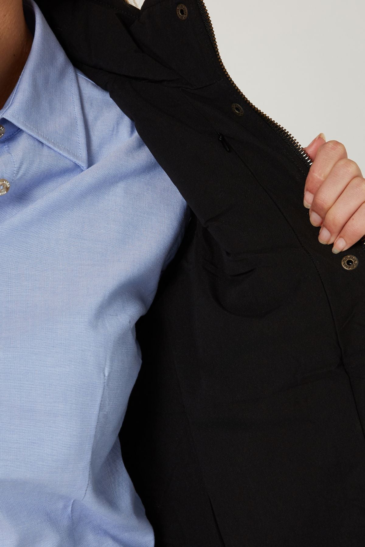 A person wearing a light blue button-up shirt is holding open the Pilbara Ladies Vest, a black winter vest with polyfill padding. Only part of their upper body and hand are visible against a plain background.