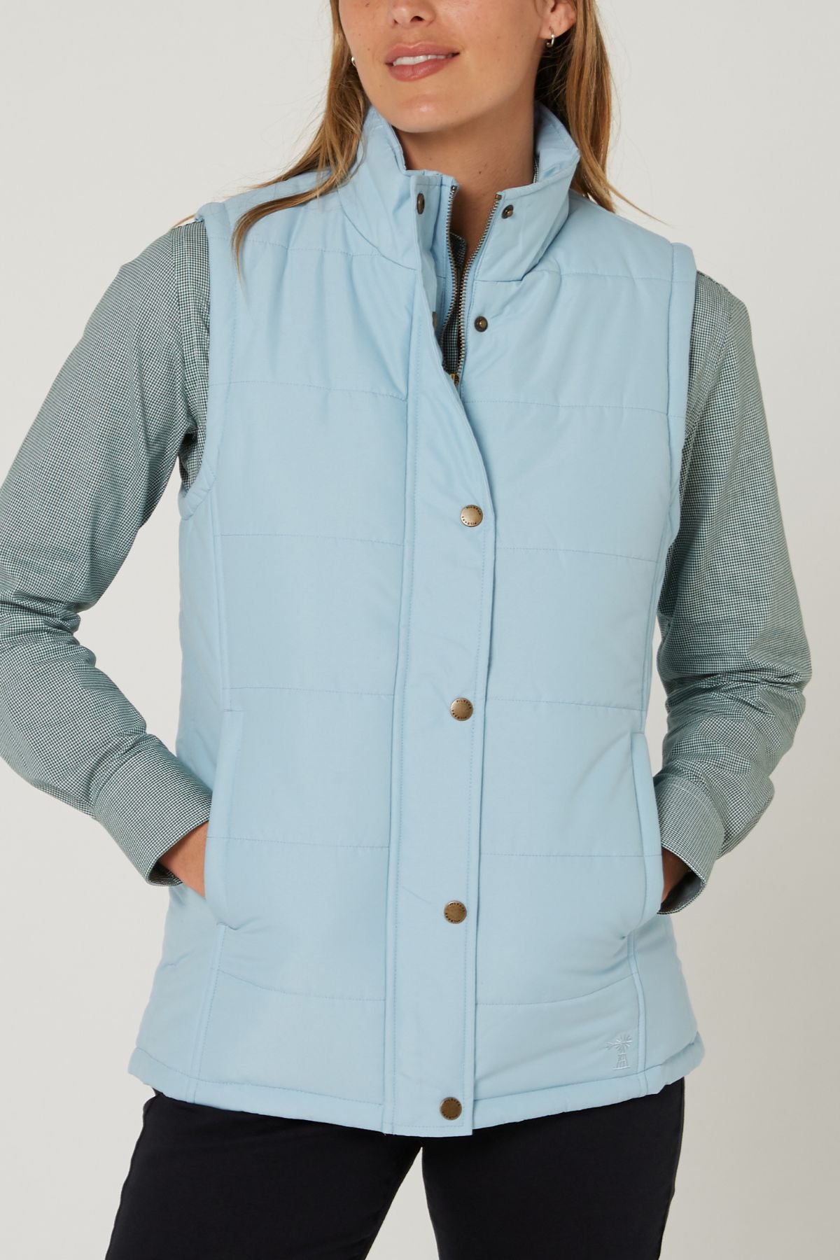 A woman wearing the Pilbara Ladies Vest in light blue with Polyfill padding over a light green shirt stands with hands in pockets against a plain background.