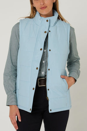 A woman wears the Pilbara Ladies Vest, a light blue microfibre waterproof winter vest with Polyfill padding, high collar, and snap buttons, over a gray-green button-up shirt and dark pants, standing with one hand in her pocket.