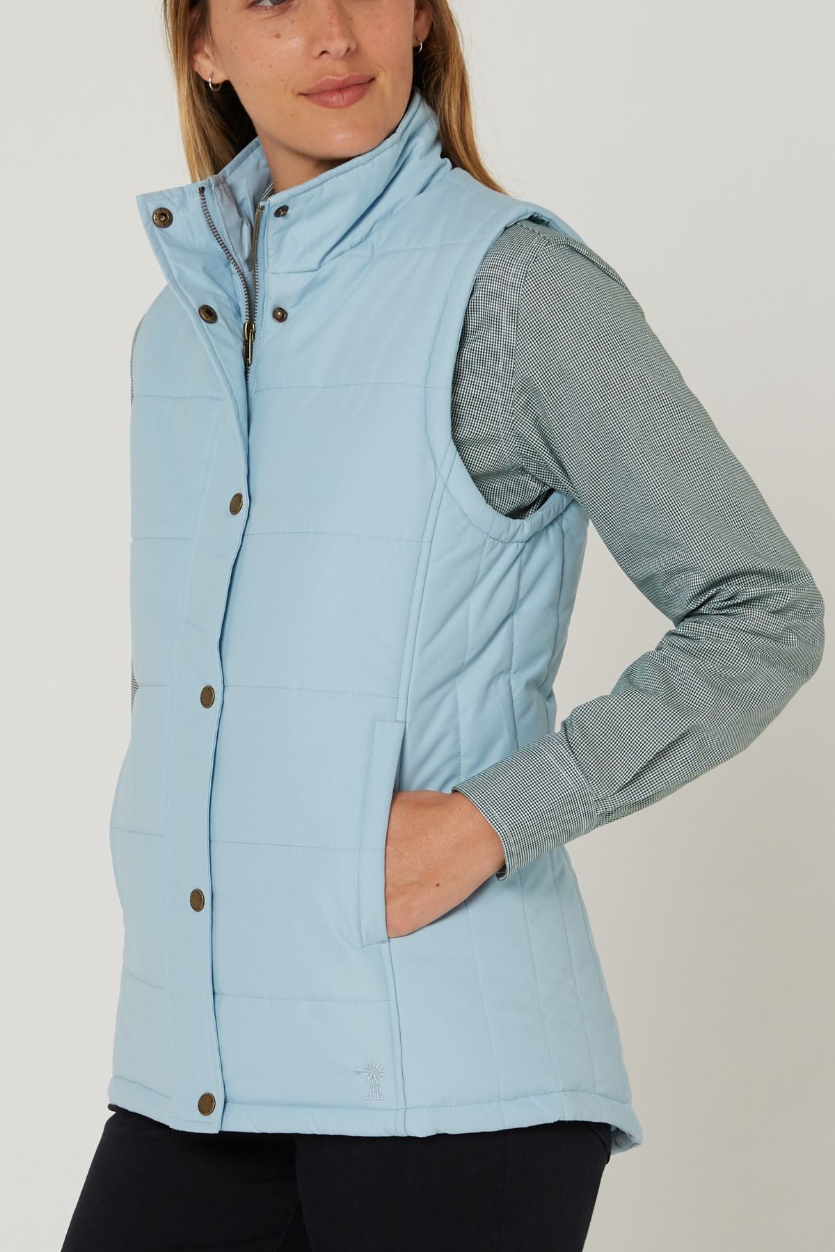 A woman wearing the Pilbara Ladies Vest in light blue with Polyfill padding over a gray-green long-sleeve shirt, one hand in her vest pocket, stands against a plain background.