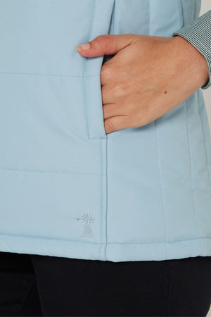 A person wearing a light blue Pilbara Ladies Vest with Polyfill padding and a small windmill logo near the hem, right hand in pocket, layered over a checkered shirt for extra warmth.