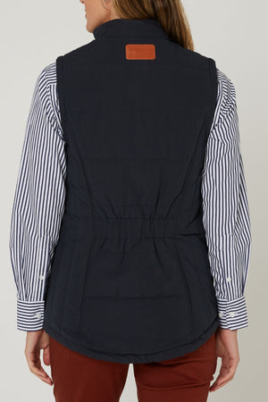 A person is shown from the back wearing the Pilbara Ladies Vest in navy blue microfibre over a white and navy striped shirt with rust-colored pants, set against a plain background.
