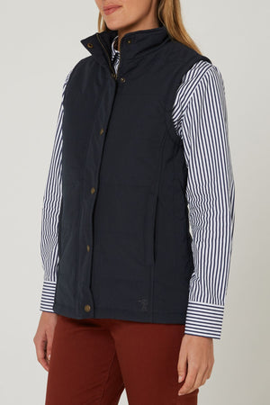 A woman wearing the Pilbara Ladies Vest in navy blue with Polyfill padding, styled over a white shirt with black stripes and rust-colored pants, stands against a plain light background.