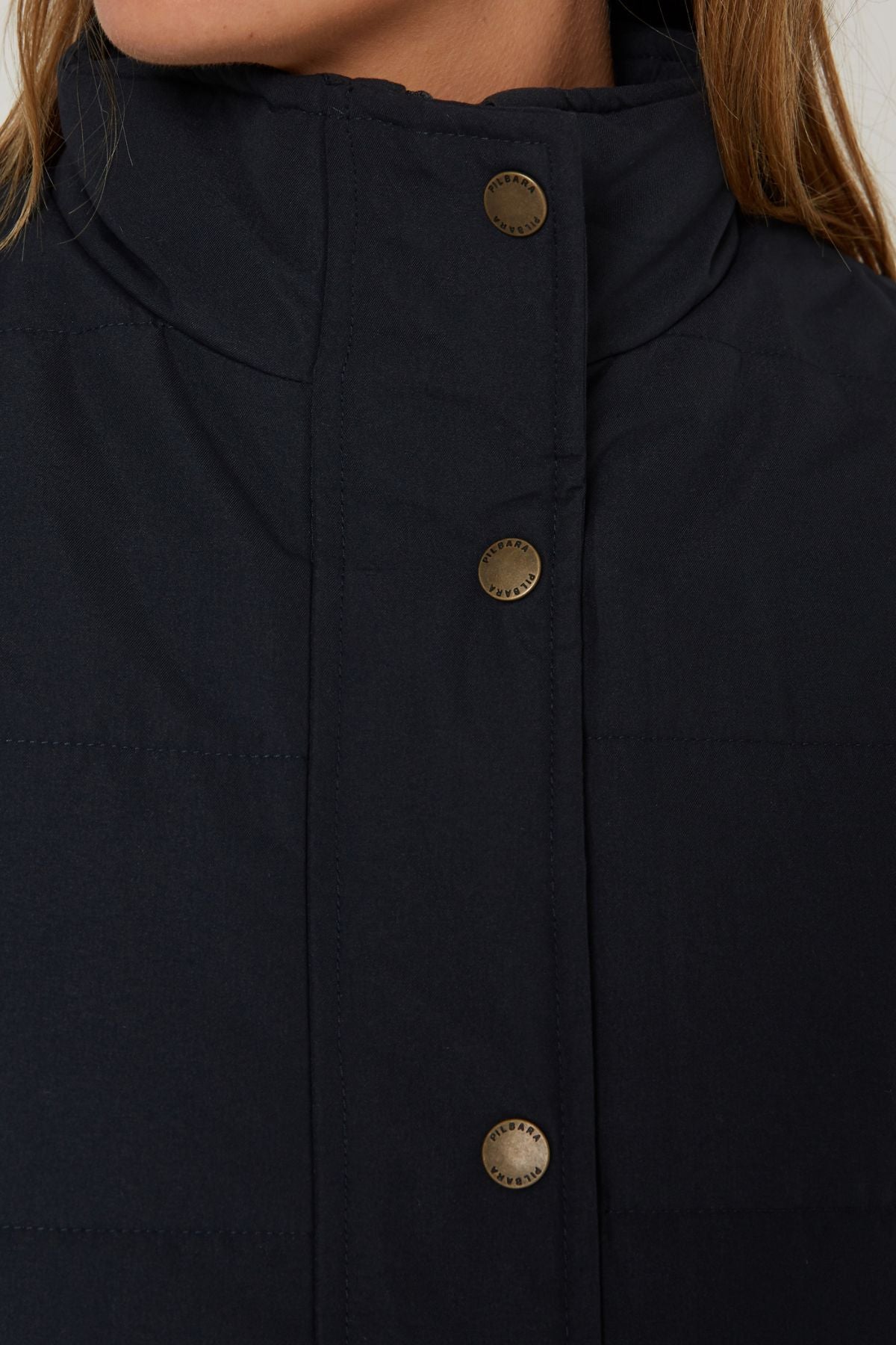 Close-up of a person wearing the Pilbara Ladies Vest, a dark quilted design with polyfill padding and metallic snap buttons. The high collar partially covers their lower face, complementing light brown hair—a cozy pick for cold weather.