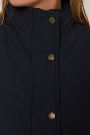 Close-up of a person wearing the Pilbara Ladies Vest, a dark quilted design with polyfill padding and metallic snap buttons. The high collar partially covers their lower face, complementing light brown hair—a cozy pick for cold weather.