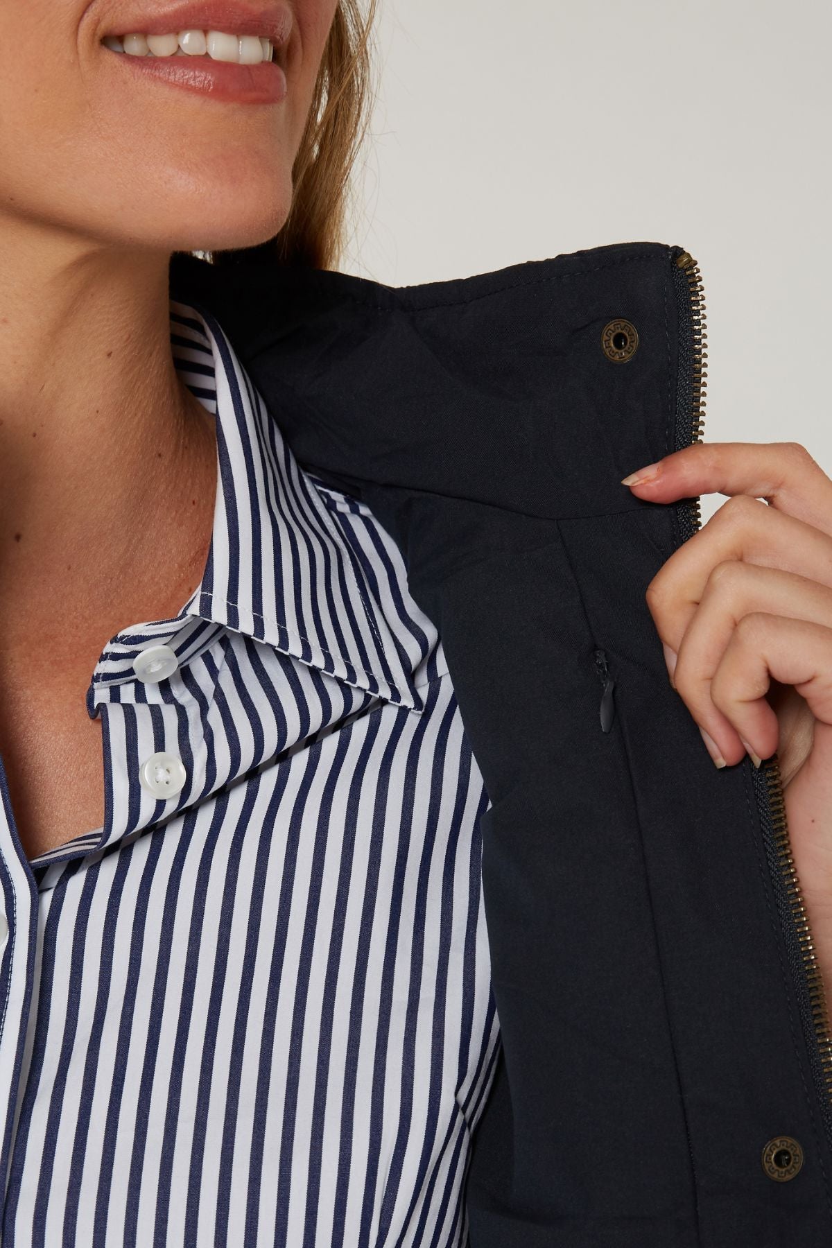 A woman smiles, showing only the lower half of her face, while wearing a blue and white striped shirt and holding open the collar of the Pilbara Ladies Vest to display its polyfill padding, zipper, and button details.