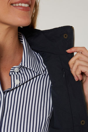 A woman smiles, showing only the lower half of her face, while wearing a blue and white striped shirt and holding open the collar of the Pilbara Ladies Vest to display its polyfill padding, zipper, and button details.