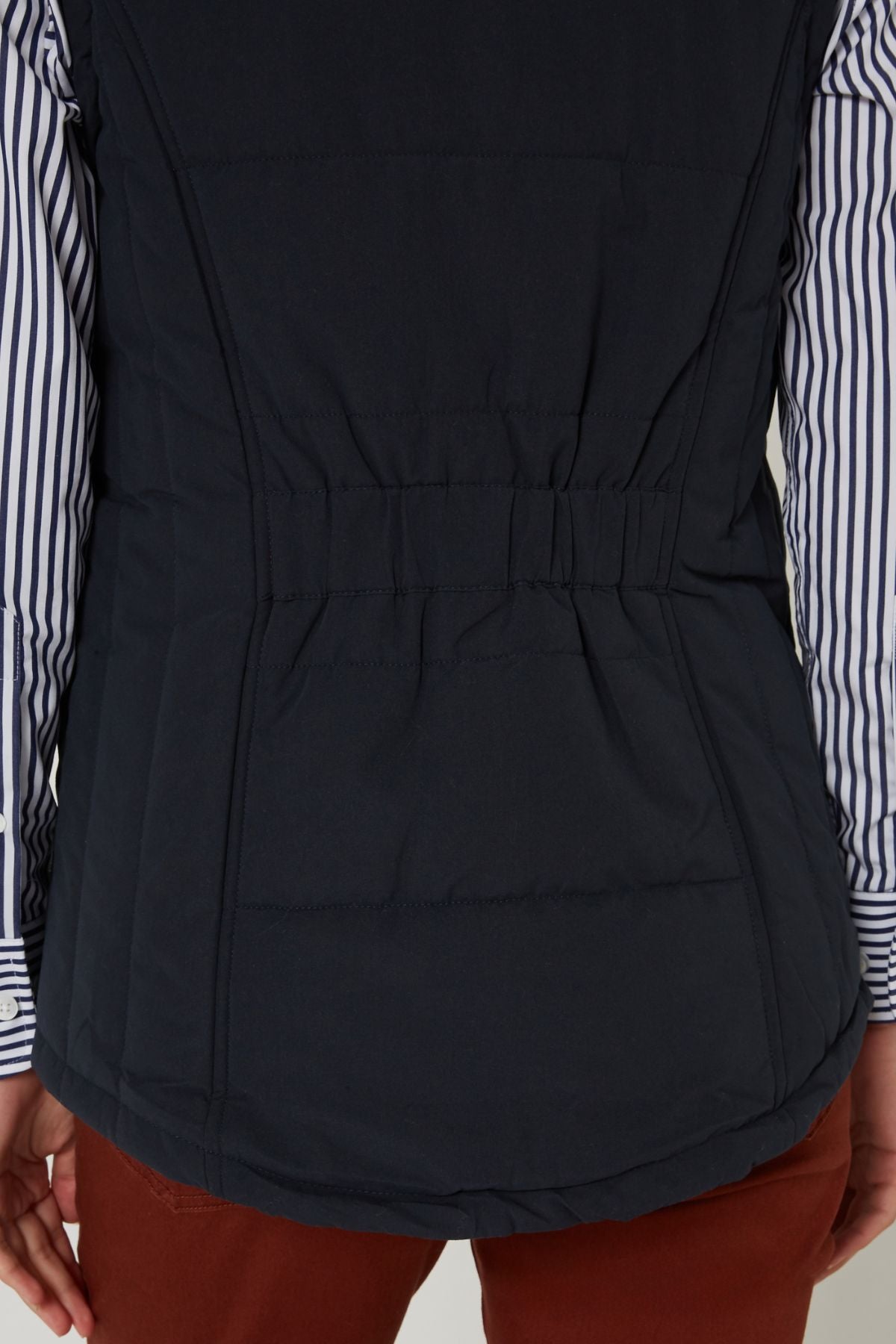 A person is seen from the back, hands relaxed at their sides, wearing the Pilbara Ladies Vest in navy with polyfill padding over a blue and white striped shirt and rust-colored pants.