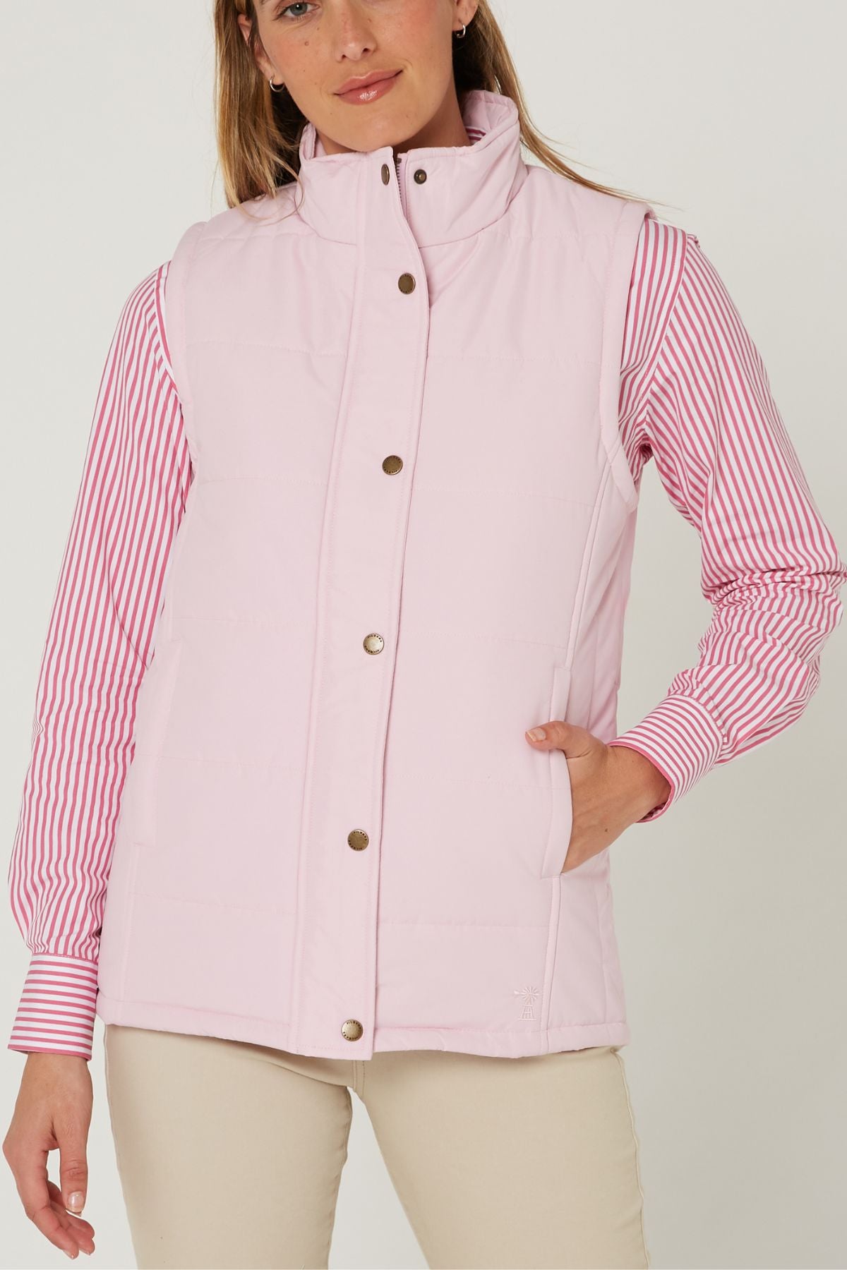 A woman wears the Pilbara Ladies Vest in light pink with Polyfill padding over a pink and white striped shirt and beige pants, standing against a plain background with one hand in her vest pocket.