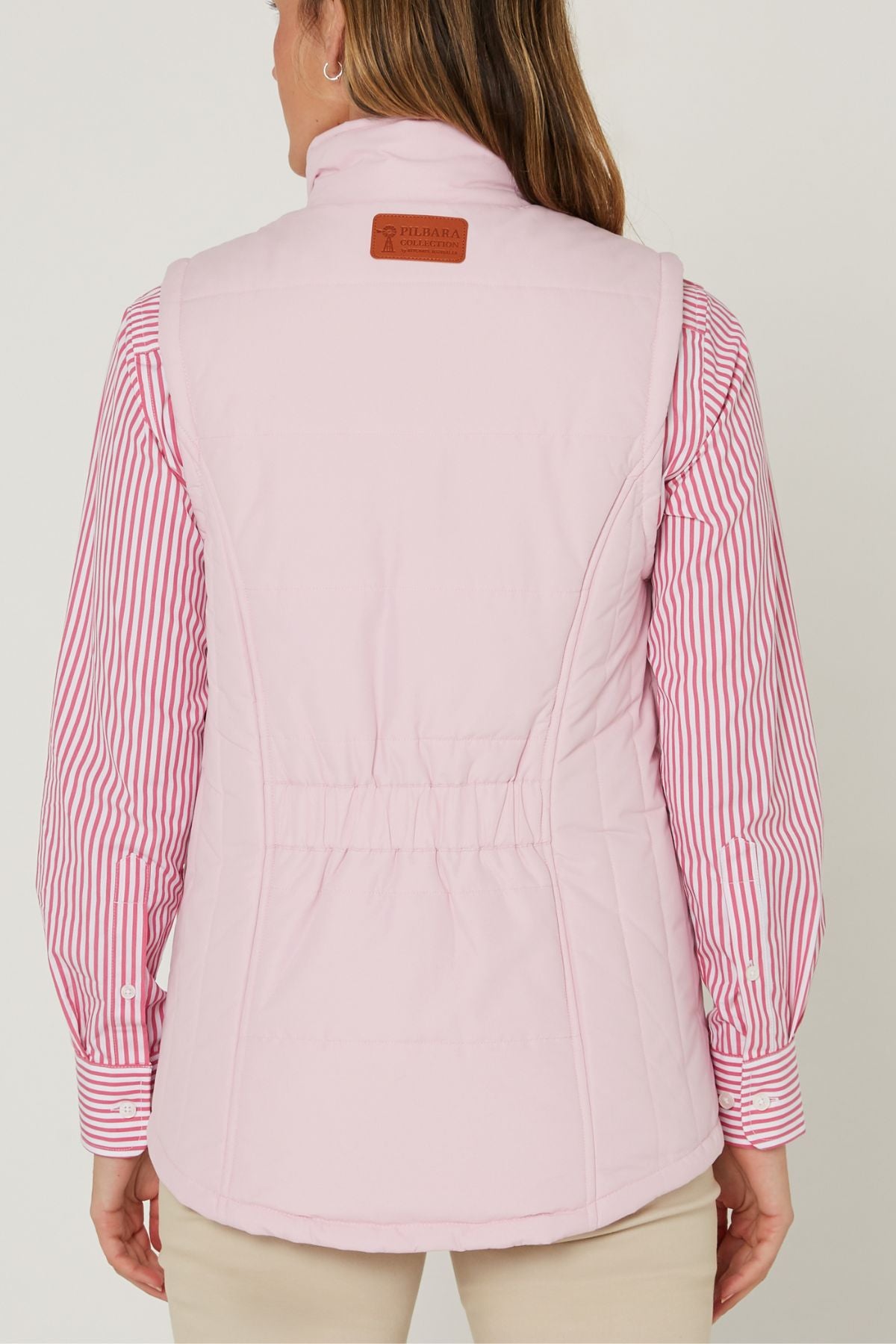 A person is seen from the back wearing the Pilbara Ladies Vest in light pink with Polyfill padding, featuring a brown leather patch near the collar and seam details, over a pink and white striped shirt and beige pants.