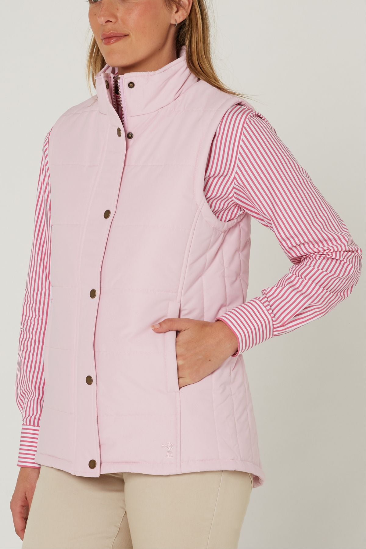 A woman models the Pilbara Ladies Vest in light pink with Polyfill padding over a pink and white striped shirt and beige pants, standing with her hand in the vest pocket against a plain white background.