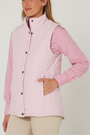 A woman models the Pilbara Ladies Vest in light pink with Polyfill padding over a pink and white striped shirt and beige pants, standing with her hand in the vest pocket against a plain white background.