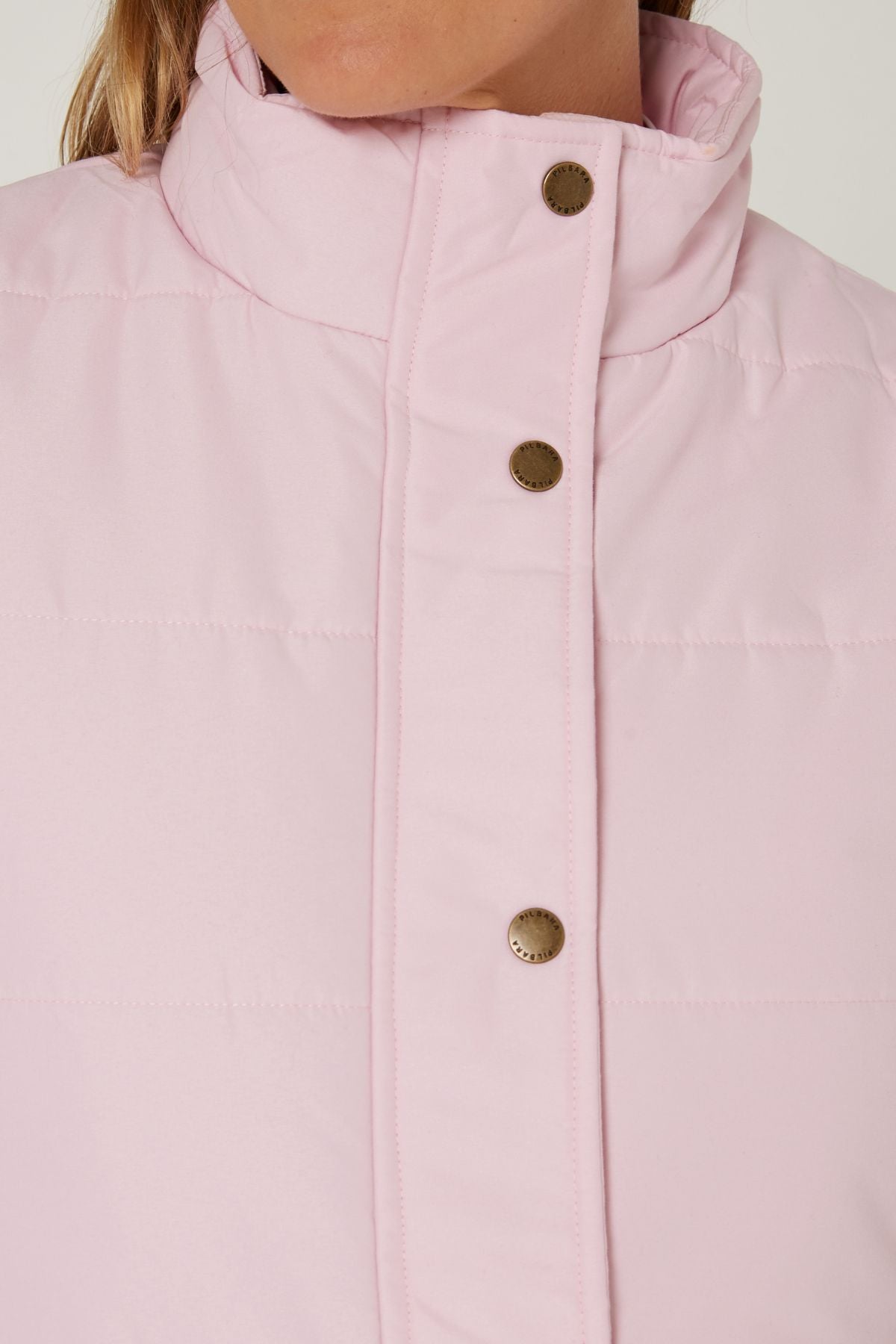 Close-up of a person wearing the Pilbara Ladies Vest—a light pink quilted vest with Polyfill padding, high collar, and metal snap buttons. The lower part of the face is partially shown.