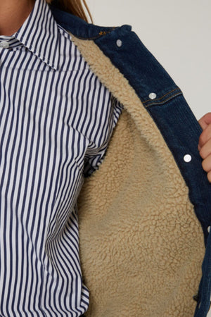 A person in a blue and white striped shirt holds open the Pilbara Lauren Sherpa Lined Jacket, showing its beige fleece lining.