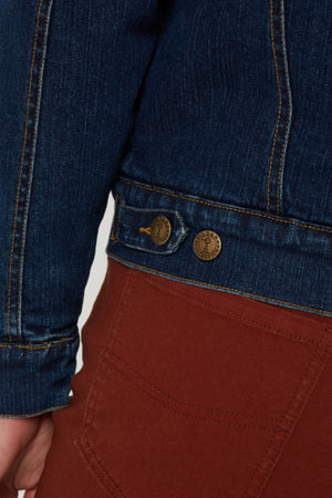 Close-up of a person wearing the Pilbara Lauren Sherpa Lined Jacket in blue denim with brass buttons and yellow stitching, paired with reddish-brown pants. The warm, casual jacket partially covers the pocket area of the pants.