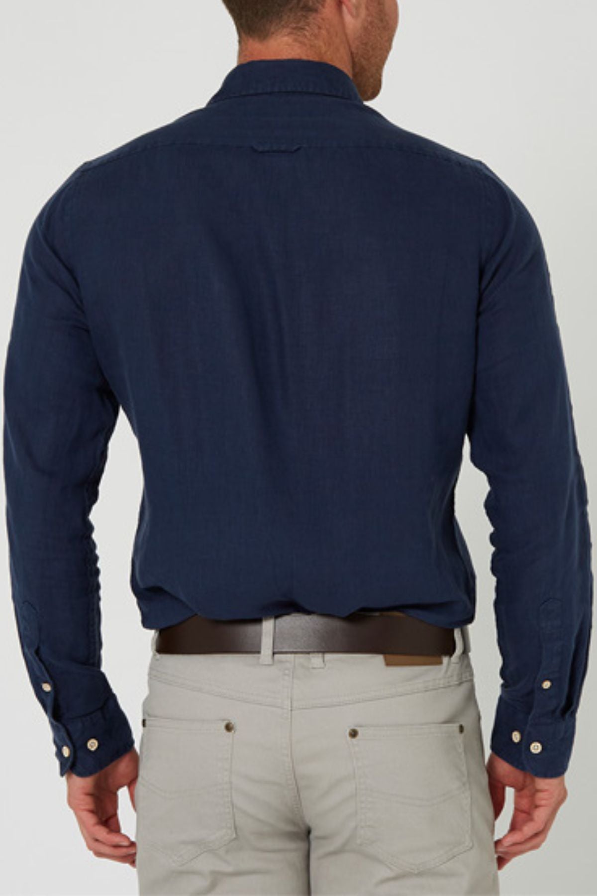 A man seen from behind wears a dark blue Pilbara Linen Shirt with buttoned cuffs and light beige trousers, standing upright against a plain light background.