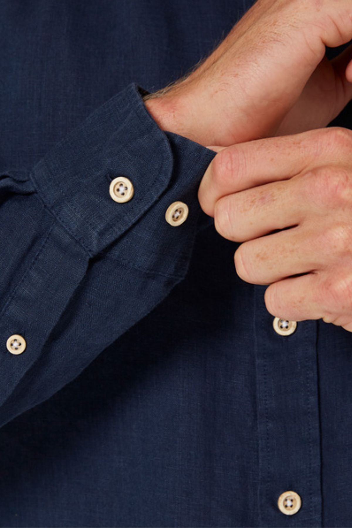 A person buttons the cuff of the Pilbara Linen Shirt in navy blue, showing only their hands and part of the long sleeve with light-colored buttons.