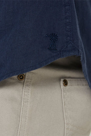 Close-up of the Pilbara Linen Shirt in navy blue, featuring a small windmill embroidery near the hem. Paired with light beige pants showcasing visible pockets and brown buttons.