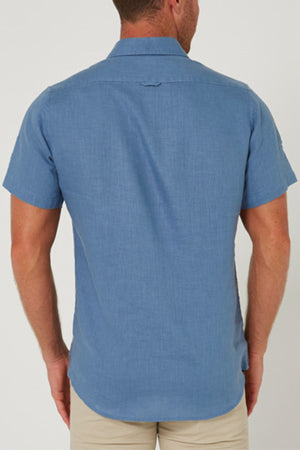A man stands with his back to the camera, wearing a Pilbara Linen Short Sleeve Shirt in light blue and beige pants against a plain white background.