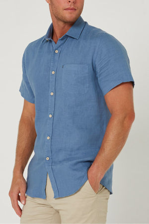 A man in the Pilbara Linen Short Sleeve Shirt in light blue, featuring a chest pocket, pairs it with beige pants and stands with one hand in his pocket against a plain background.