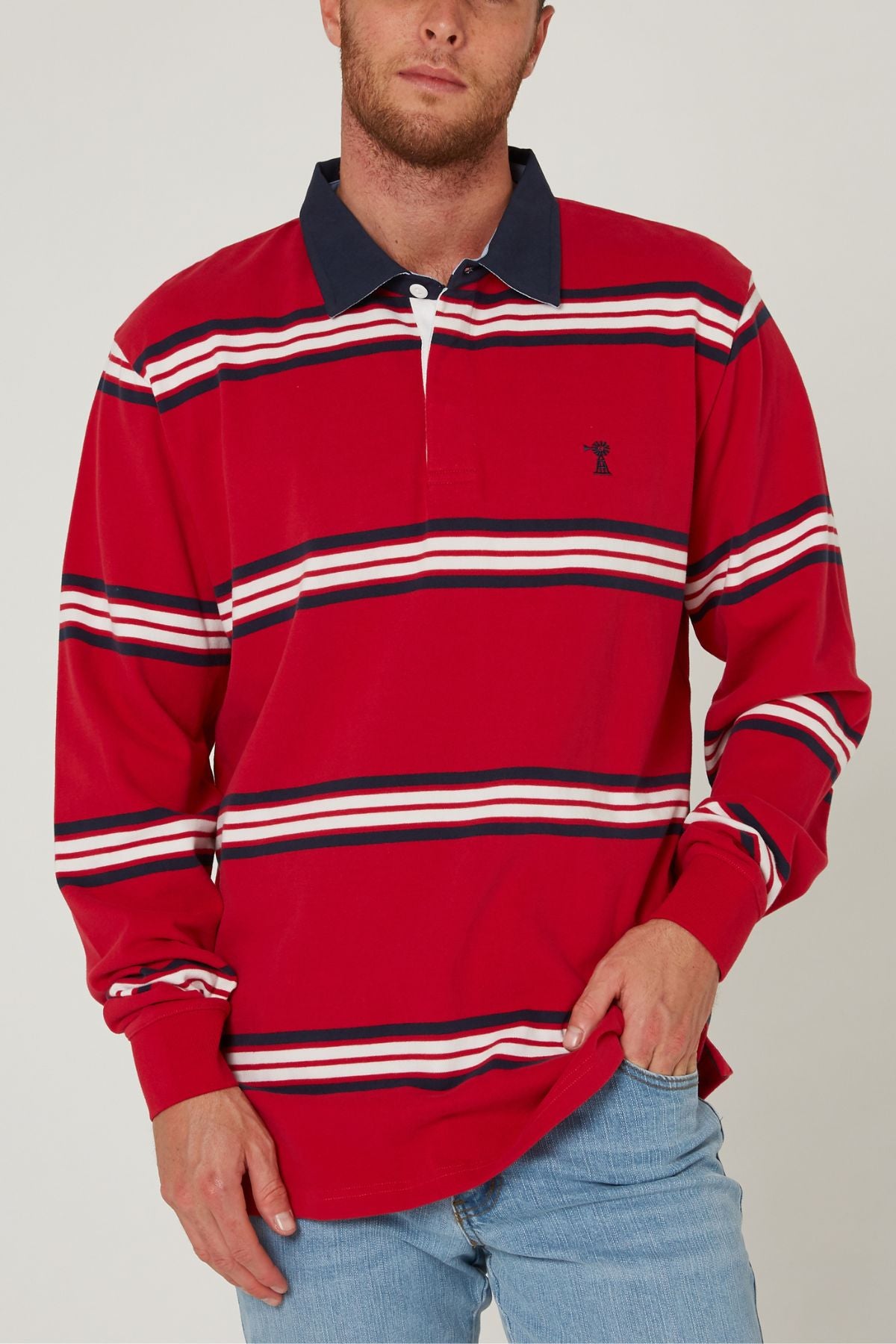 A man wearing a long-sleeve Pilbara Rugby Jersey with red, white, and navy stripes, a navy collar, and light blue jeans stands with one hand in his pocket.