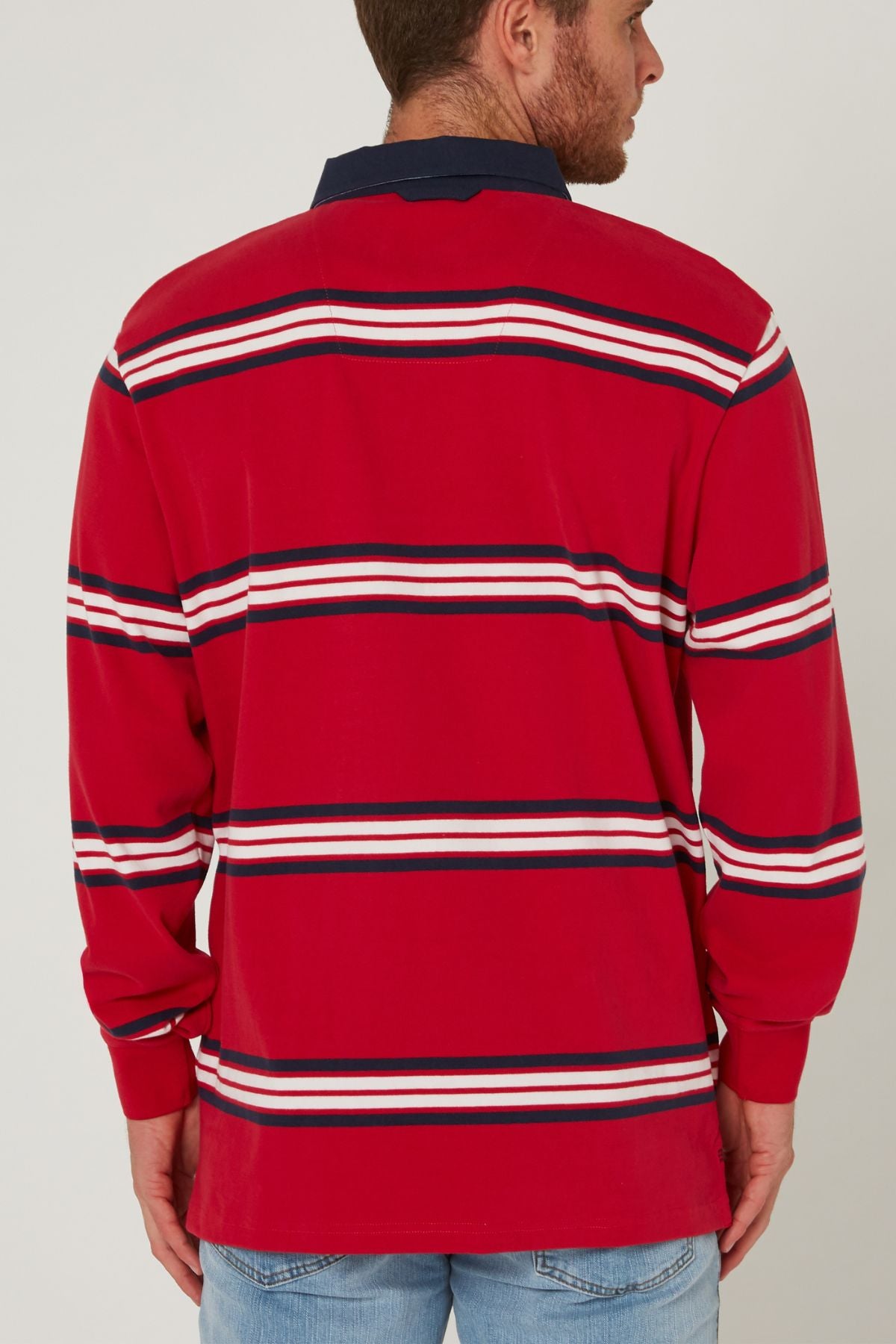 A man faces away wearing the Pilbara Rugby Jersey, featuring red with horizontal white, navy, and black stripes, a navy collar, and paired with light blue jeans.