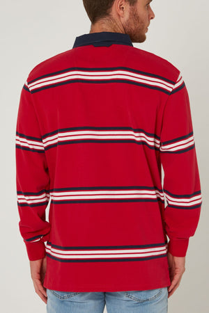 A man faces away wearing the Pilbara Rugby Jersey, featuring red with horizontal white, navy, and black stripes, a navy collar, and paired with light blue jeans.
