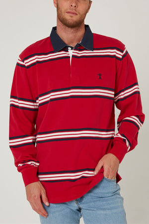 A man wearing a long-sleeve Pilbara Rugby Jersey with red, white, and navy stripes, a navy collar, and light blue jeans stands with one hand in his pocket.