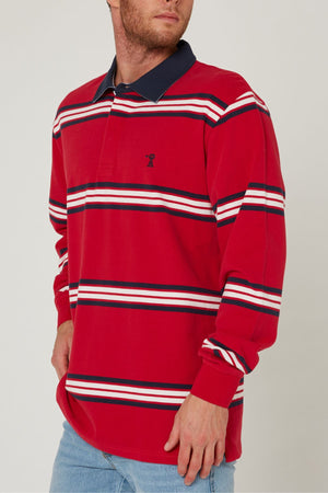 A man in a red Pilbara Rugby Jersey with white, navy, and black stripes and a navy collar—100% cotton—stands with one hand on his hip, wearing light blue jeans and facing slightly away from the camera.