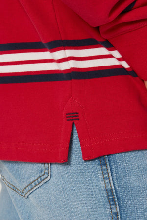 A close-up of a person in light blue jeans and a Pilbara Rugby Jersey, made from 100% cotton, in red with white and navy horizontal stripes, featuring a small side slit with extra navy blue stitching.