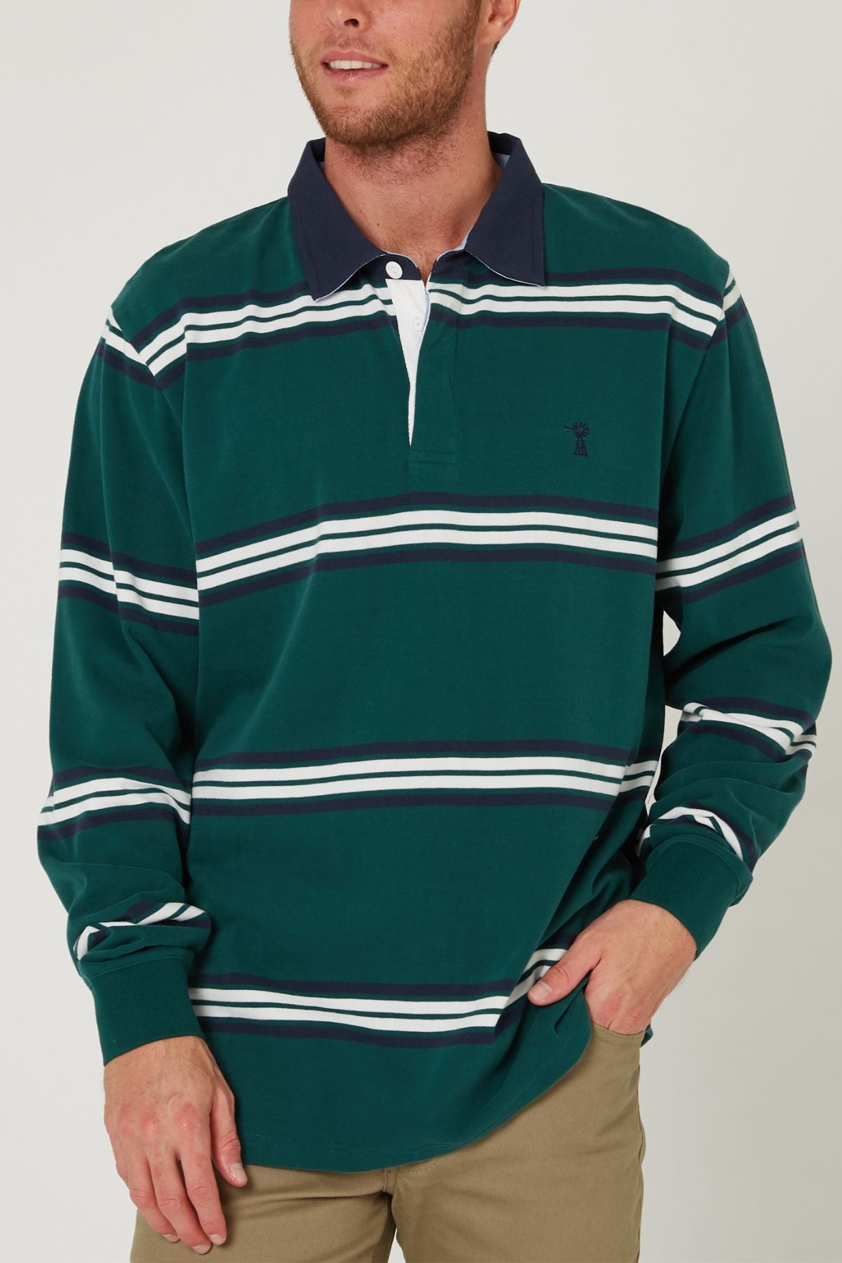 A man wearing the Pilbara Rugby Jersey—green 100% cotton with white and navy stripes—and khaki pants stands with one hand in his pocket, smiling slightly.
