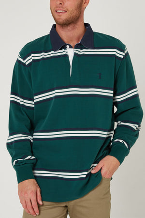 A man wearing the Pilbara Rugby Jersey—green 100% cotton with white and navy stripes—and khaki pants stands with one hand in his pocket, smiling slightly.