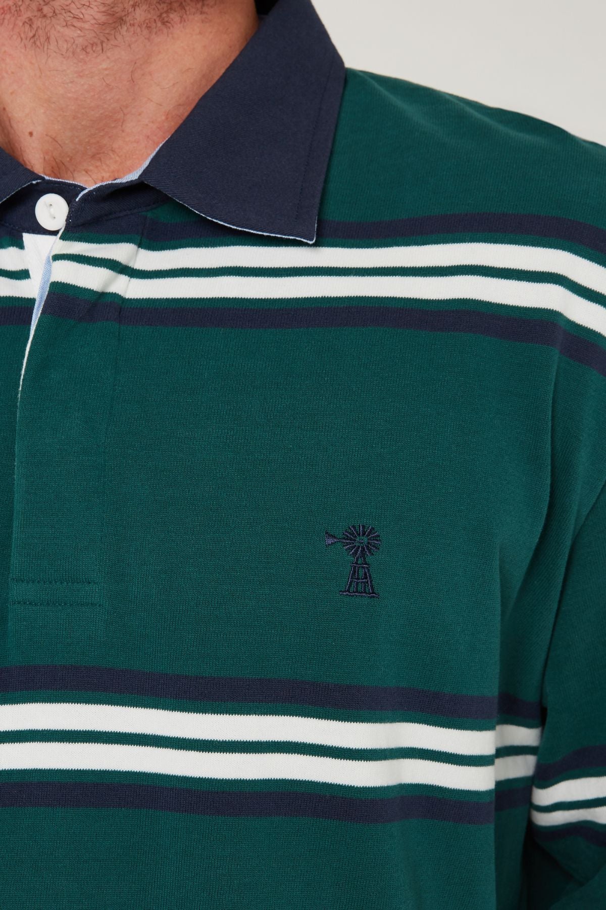 Close-up of a person wearing the Pilbara Rugby Jersey, featuring green with navy and white horizontal stripes, a navy collar, and an embroidered windmill Pilbara logo on the chest. Made from 100% cotton for comfort.
