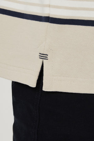 Close-up of the hem of the Pilbara Rugby Jersey, a beige shirt with dark blue stripes, classic rugby style, featuring a small side slit and decorative stitching, worn over dark pants.