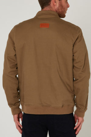 A man is seen from behind wearing a tan Pilbara Sherpa Bomber Jacket with a small rectangular patch on the upper back and dark pants, the sherpa-lined bomber standing out against a plain, light-colored background.