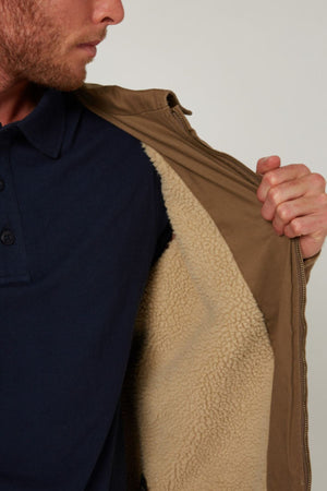 A man in a navy blue polo shirt holds open the Pilbara Sherpa Bomber Jacket, revealing its light beige sherpa-lined interior made of cotton stretch twill. The image is cropped to show just the lower half of his face and upper torso.
